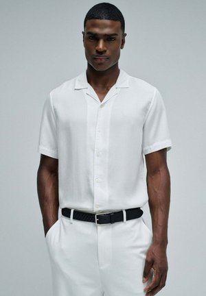 Joven con camisa blanca de manga corta abotonada y pantalones, cinturón negro texturizado, de pie con las manos en los bolsillos frente a un fondo sencillo.