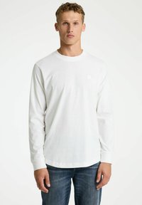Weißes Langarmshirt aus Baumwolle. Runder Ausschnitt, ein kleines quadratisches Logo auf der linken Brust und gerader Saum. Getragen mit blauen Jeans.