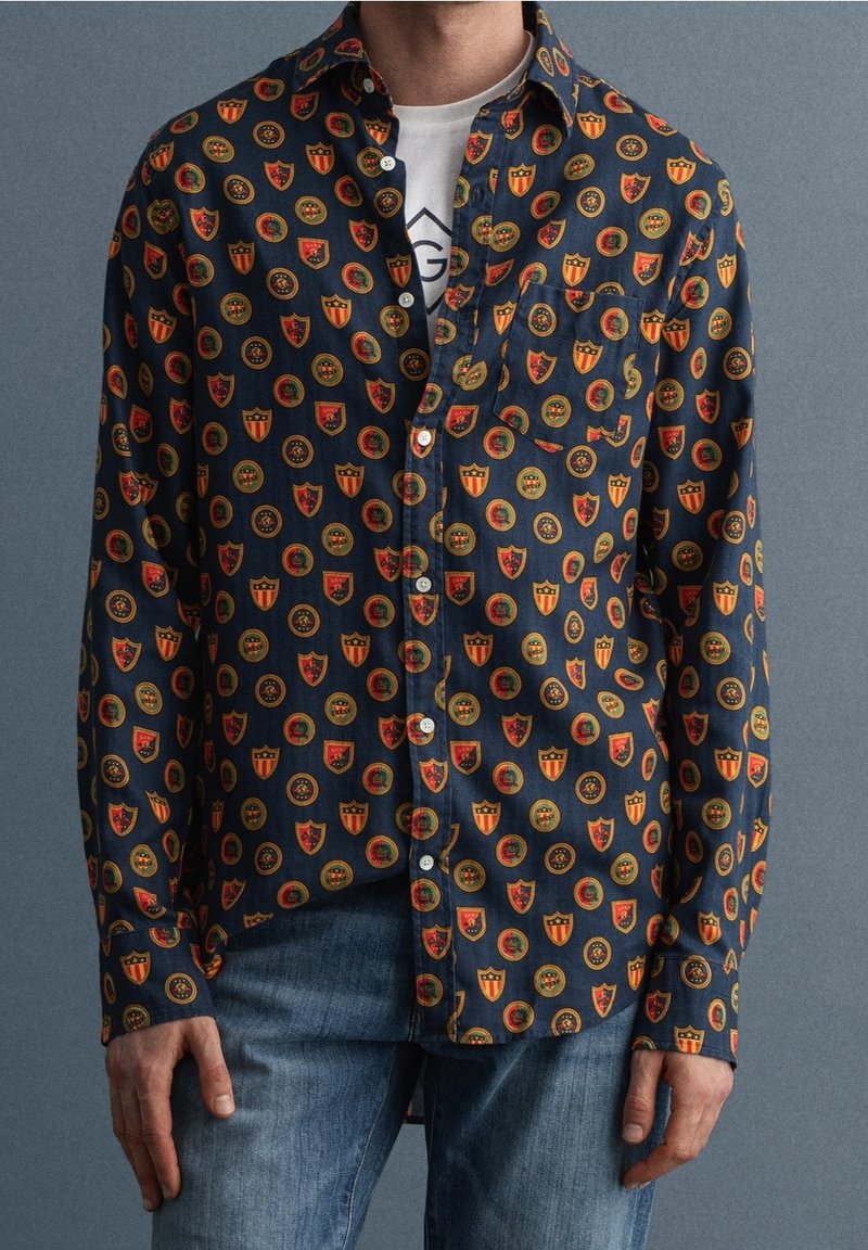 Chemise pour homme à manches longues en tissu foncé, ornée d'un motif coloré de boucliers et de cercles. Devant boutonné avec une poche poitrine unique.