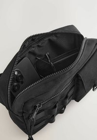 Black fabric bag with a zipper, partially open to reveal a black smartphone and sunglasses inside, featuring a smooth texture and straps.