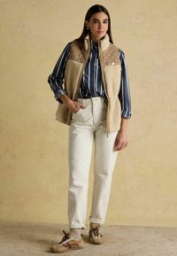 Beežist fliisist ja trapetsikujulisest vestist sinise ja valge triibulise särgi peal, kombineeritud kõrge vöökoha valgete teksade ja beežide tossudega. Neutraalne taust.