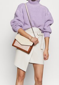 Violetti neulepaita, vaalean kerman ja vaaleanpunaisen väri-injektoitu olkalaukku ruskeilla yksityiskohdilla. Valkoinen kietaisuhame, jossa on solmukiinnitys, sileä pinta.