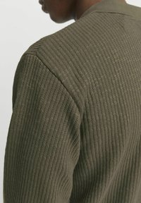 Jersey de color verde oliva con textura, presenta un cuello redondeado y mangas ajustadas, mostrando un primer plano del área del hombro.