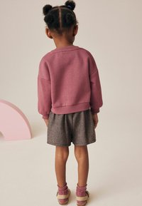 El atuendo de la niña incluye una sudadera rosa con escote redondo y mangas abullonadas, combinada con pantalones cortos de estampado de hormiguita grises y sandalias a juego.