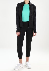 Giacca sportiva nera, top aderente turchese, leggings neri e scarpe da ginnastica bianche. La giacca ha una zip; l'outfit enfatizza un look sportivo ed elegante.