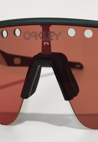 Rot getönte Sonnenbrille mit einer geschwungenen Linse, schwarzen Akzenten und auffälligen Ausschnitten. Das Logo "OAKLEY" ist auf der Linse angebracht.