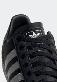 Czarny sneaker Adidas z czarnymi sznurowadłami, trzema białymi paskami po boku oraz logo Adidas trefoil na języku.