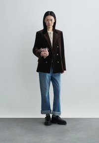 Brown velvet blazer with notched lapels, paired with light blue cuffed jeans and black chunky shoes. Holding a small, pale pink bag.