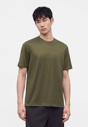 Jeune homme aux cheveux courts noirs portant un T-shirt vert olive avec un logo d'aigle en relief et un pantalon noir, debout devant un fond blanc uni.