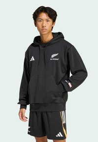 Schwarzer Hoodie mit Fronttasche, weißem "ALL BLACKS"-Logo und Adidas-Streifen; kombiniert mit schwarzen Shorts mit orangefarbenen Akzenten und Logos.