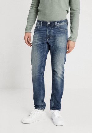 Mann trägt verblasste blaue Jeans, weiße Turnschuhe und ein langärmeliges hellgrünes Hemd, steht vor einem schlichten weißen Hintergrund.