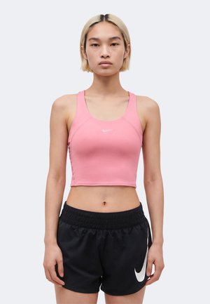 Młoda osoba z krótkimi blond włosami, ubrana w różowy krótki top Nike i czarne sportowe spodenki Nike, stoi zwrócona twarzą do przodu.