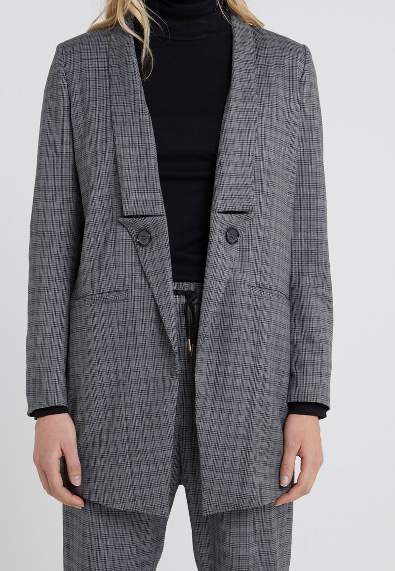 Femme portant un blazer gris à carreaux et un pantalon assorti par-dessus un pull à col roulé noir, sur un fond uni.