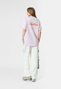 Camiseta extragrande de color rosa claro con "Sin restricciones" en texto naranja en la parte posterior. Combinada con pantalones blancos de corte holgado y zapatos de eslora grises.
