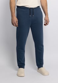Dunkelblaue Jogginghosen mit elastischem Bund, Kordelzug, glatter Textur und schmal zulaufenden Beinen. Kombiniert mit hellbeigen Slippers.