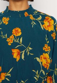 Teal klänning med blommönster i gult och orange, med en smockad halsringning och mjuk tygtextur.