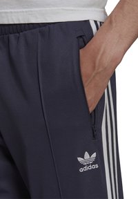 Pantaloni sportivi blu navy realizzati in tessuto liscio, con strisce bianche sui lati, una tasca con zip e un logo ricamato sul davanti.