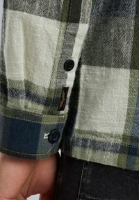 Chemise à carreaux verte et blanche avec des poignets à boutons, présentant un tissu texturé, des carreaux bleu marine contrastants et une étiquette de logo sur l'ourlet.