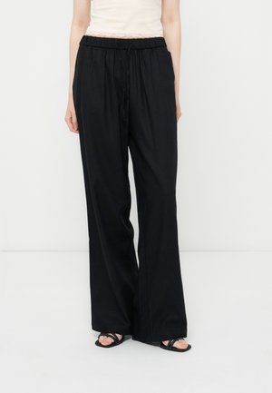 Γυναίκα που φοράει φαρδιά μαύρη παντελόνα με κορδόνι και μαύρα σανδάλια με ανοιχτά δάχτυλα σε απλό λευκό φόντο.