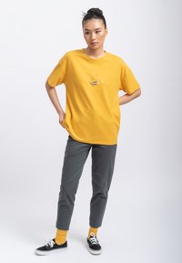 Gele casual t-shirt met korte mouwen met abstract ontwerp. Gecombineerd met grijze, tapered broek en zwarte sneakers.