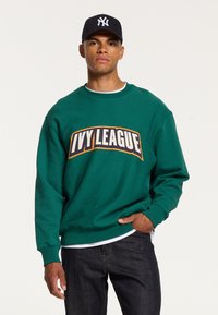 Felpa verde con colletto rotondo con "IVY LEAGUE" stampato in lettere in grassetto. Abbinata a jeans scuri, il modello indossa anche un berretto da baseball nero.