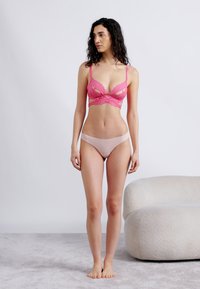 Γυναίκα με μακριά σκούρα μαλλιά, που φοράει ροζ δαντελένιο σουτιέν και γυμνό χρώμα εσώρουχα, σκυφτή δίπλα σε λευκή στρογγυλή πολυθρόνα, χωρίς παπούτσια.