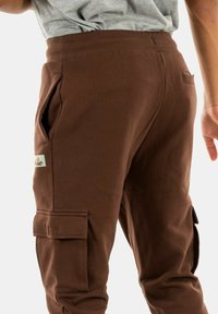 Pantalones cargo marrones con bolsillos laterales, cinturilla elástica y detalles en parches. Fabricados con un tejido suave de textura lisa.