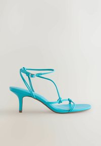 Sandale en cuir bleu à brides avec un petit talon kitten et une fermeture à boucle à la cheville sur fond neutre.