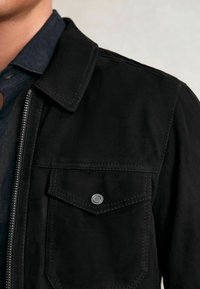 Melna zamšā jaka ar rāvējslēdzēja aizdari, norādītu apkakli, kreiso krūšu kabatu un tonālu šuvumu. Mīksta tekstūra, minimāla aparatūras detaļa.