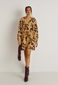 Φόρεμα κοντομάνικο σε μπεζ χρώμα με σκούρες καφέ αφηρημένες σχέσεις, cinched στη μέση με κορδόνι, συνδυασμένο με τσάντα σε σκούρο μωβ και μπότες.