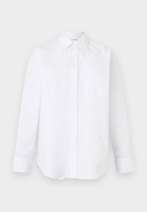 Femme aux cheveux noirs et raides portant des lunettes, une chemise blanche oversize, un pantalon large foncé et des chaussures bordeaux, debout contre un mur gris uni.