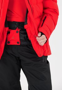 Czerwona kurtka puchowa z zamkiem błyskawicznym i akcentami guzika, noszona na czerwonym topie, w połączeniu z czarnymi spodniami z elastyczną talią i bocznym zamkiem.