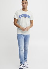 Gai pelēks kokvilnas T-krekls ar uzrakstu "UNIVERSITY" zilās arkas formā, valkāts kopā ar gaiši ziliem džinsiem un melnbaltiem sporta apaviem.