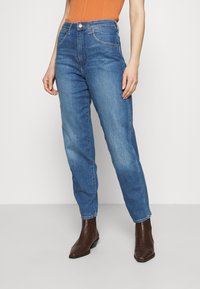 Džíny s vysokým pasem a rovnými nohavicemi v modré barvě s vyšisovaným vzhledem, obsahující přední kapsy a kovové zapínání na knoflík. Spárováno s hnědými kotníkovými botami.