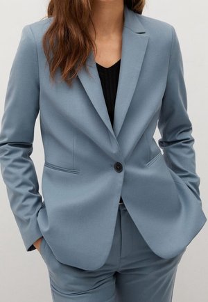 Femme portant un blazer bleu clair cintré et un pantalon assorti, les mains dans les poches, sur un haut noir.