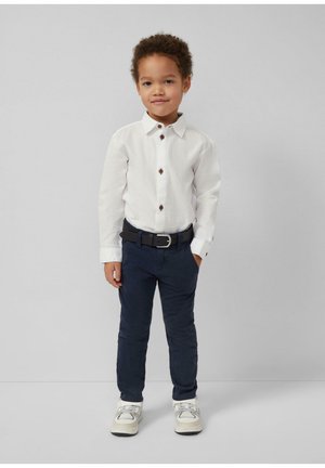 Małe dziecko stojące, ubrane w białą koszulę zapinaną na guziki, ciemne spodnie z czarnym paskiem oraz białe trampki na jednolitym szarym tle.