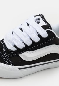 Adidași negri din pânză cu șireturi albe, având o dungi albă pe laterale, cusături negre și talpă din cauciuc texturat. Vedere de aproape care evidențiază detaliile.