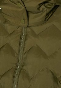 Gros plan sur un tissu matelassé vert olive avec une fermeture éclair et des boutons-pression, présentant un motif de coutures en chevron.