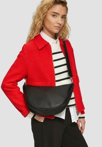 Schwarze Leder-Schultertasche mit geschwungenem Design, strukturiertem Oberflächenmaterial und einem einzigen verstellbaren Tragegurt. Wird zu einer roten Jacke getragen.
