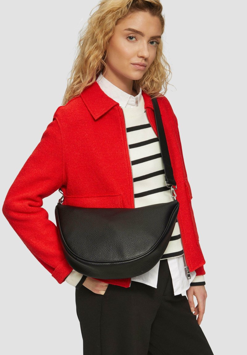 Schwarze Leder-Schultertasche mit geschwungenem Design, strukturiertem Oberflächenmaterial und einem einzigen verstellbaren Tragegurt. Wird zu einer roten Jacke getragen.