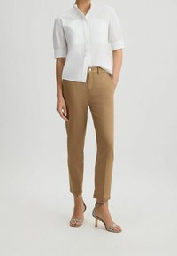 Femme portant une chemise blanche à manches courtes boutonnée, un pantalon beige à la cheville et des sandales à talons hauts au motif serpent.