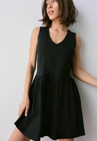 Schwarzes ärmelloses Kleid mit V-Ausschnitt, tailliertem Oberteil und schwingendem Rock. Hergestellt aus leichtem Material, das schlichte, klare Linien präsentiert.