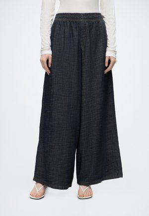 Weit geschnittene dunkelblaue Jeanshose mit elastischem Bund, getragen zu weißem Langarmshirt und weißen offenen Sandalen.
