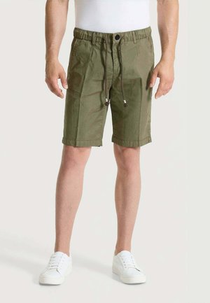 Uomo con pantaloncini verde oliva fino al ginocchio con vita regolabile con coulisse e sneakers bianche, in piedi davanti a uno sfondo semplice.