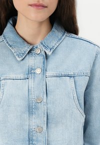 Veste en denim bleu clair avec un col classique, des boutons-pression argentés et deux poches avant avec des détails de couture.