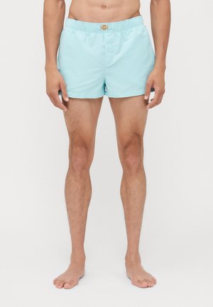Bañador de color azul claro con cinturilla elástica y detalles de hardware dorado. La tela parece suave; los pantalones cortos tienen un ajuste holgado.