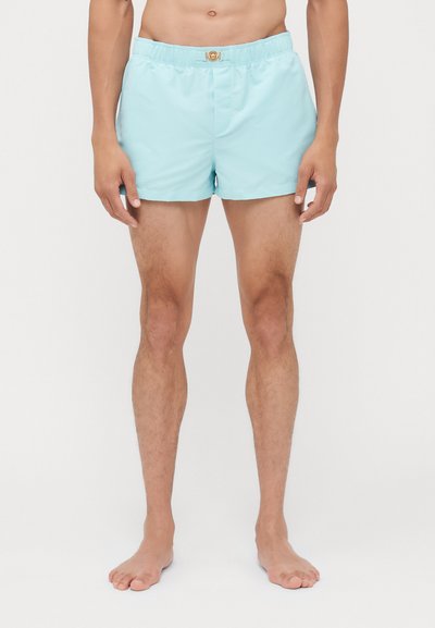 Lichtblauwe zwembroek met een elastische tailleband en details van goudkleurig metaal. De stof lijkt soepel; de shorts hebben een losse pasvorm.