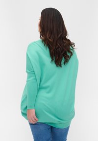 Žena s dlouhými hnědými vlasy, oblečená v volném světle zeleném svetru a modrých džínách, obrácená zády k jednoduchému bílému pozadí.