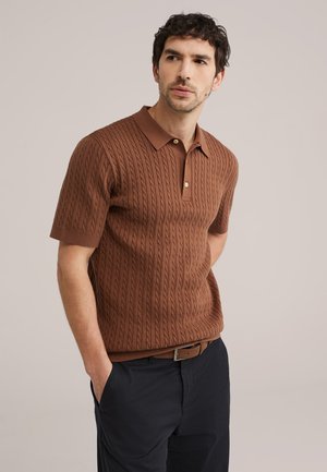 Moški s temnimi kodrastimi lasmi, oblečen v rjavo polo majico s kratkimi rokavi in pletenim vzorcem ter temne hlače, roke v žepih, nevtralno ozadje.