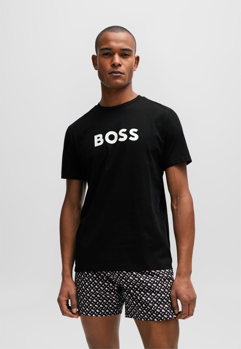 Schwarzes Baumwoll-T-Shirt mit "BOSS" in fetter, weißer Schrift, kombiniert mit gemusterten Shorts, die ein geometrisches Design in Blau-, Weiß- und Orangetönen aufweisen.